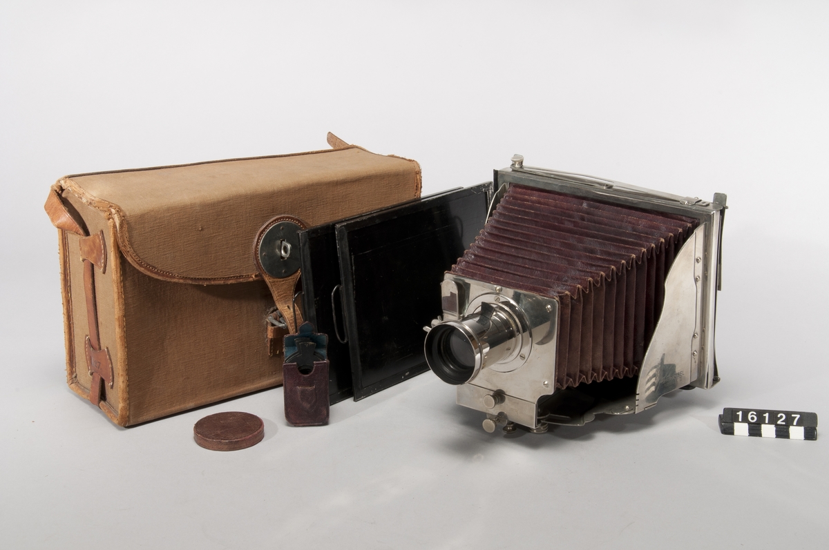 The first foldable metal camera, Model II. Constructed with a patent by H. Mader from 1888-1890. The camera was donated to the museum in January 1939.
The camera is marked with a "5" on the bellows' folds and with a circular mark bearing the letters G M R and two sword-like symbols.
The lens is marked "Aplanal Invincible" and has "VI" engraved on the back near the threads. The camera comes with 4 cassettes, 5 insert apertures, and a fabric-covered case.