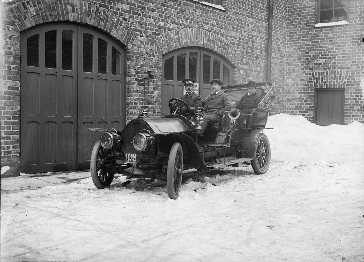 Två män med chaufför i bil utanför automobil/vagnfabrik. Troligen Gustaf LM Ericssons Automobilfabrik.