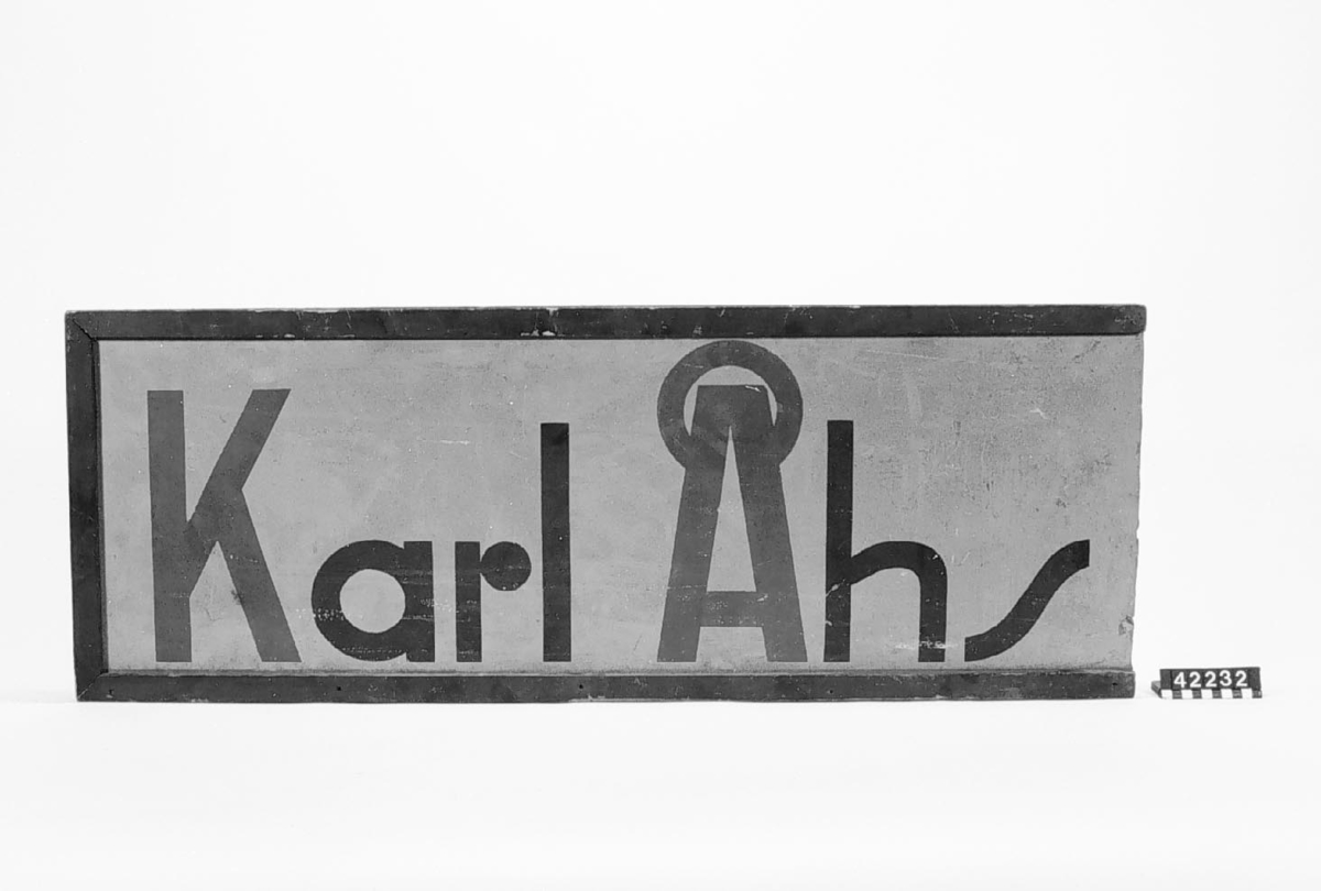 Skylt av masonit med trälister runt. Text: Karl Åhs med svarta och röda bokstäver på gul botten. Smutsig, en kortsidelist saknas.