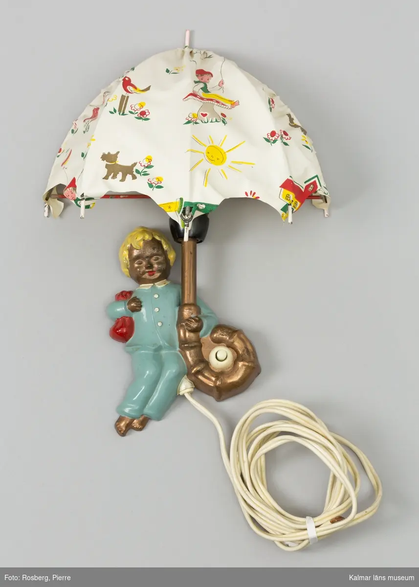 KLM 45121:1, Lampett, av metall och plast. Elektrifierad lampett för barnkammare. Guldlockigt barn i blå pyjamas håller ett paraply som utgör lampskärmen. Paraplyet har mönster av lekande barn, blommor, hundar m m. Lampetten har sladd och glödlampa men stickkontakten saknas.