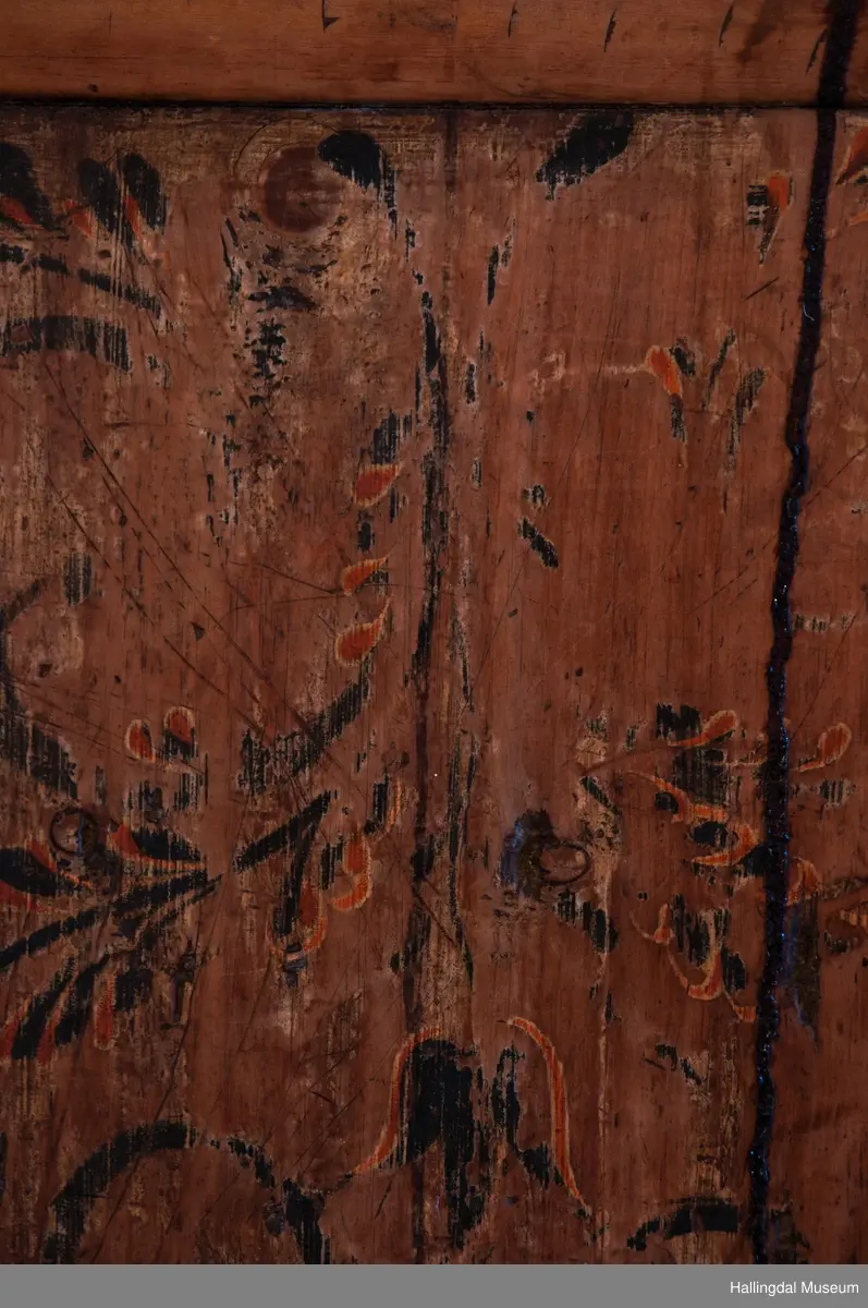 Rester etter rød og blå maling.  Gesims, skråner ned bakover. Profilert. 6 sirkulære hull i arkitraven.  Mellom disse er malt med sorte bokstaver.  Gavlene har utskårne "hylleknekter".  Bakveggen består av fire vertikale planker.  Skjøtene er dekket med profilerte lister.  Bare to biter i nederste del til venstre er fremdeles på plass.  Nederste del er lukket skap.  Tredelt, midterste del er skapdør.  Platen har profillist.  Feltene har speilfyllinger.  Blåmalte.  Klosset marmorering.  Speilfyllingenes form er som en liggen H.  Har profillist.  Skapet er satt på en ny grønmalt plate.  På døren er malt hvite bokstaver.  Nederst på døren er skåret inn bokstaver, opp-ned.  To nøkkelhull m. jernbeslag på venstre side av døren.  Over døren til venstre, en dreibar trekloss, holder igjen.  Innvendig har døren merke etter stort jernbeslag på samme side som nøkkelhullene.  To lister skåret i treverket minner om lange jernbeslag,-smalner mot nøkkelhullene.  Akantusranker i rødt, blått og hvitt, meget avslitt.  En hylle i skapet. Mangler bakvegg.  Karmen har på venstre side spor etter to jernhengsler.  Øverst på høyre kortside, mangler gesimsen en bit, og holdes til sideveggen med en jernkrampe.  Jernkrampe også på venstre side.  Skapplaten mangler list på høyre side.  Venstre side, fra gesimsen og ned har spor etter å ha stått inntil en vegg eller et annet møbel.