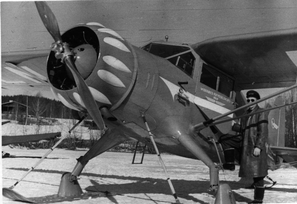 Landskap. Et fly, Hønningstad C5 Polar med skiunderstell, parkert på en snøkledd slette. Ved siden av flyet står en person i uniform, pilot/flyger.