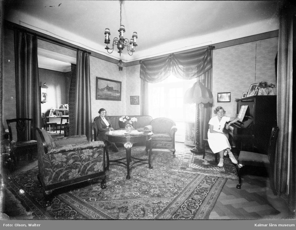En interiörbild med två kvinnor varav en sitter vid ett piano. Enligt Walter Olsons journal är bilden beställd av Fru Hilda Olsson ifrån Södra vägen 16.