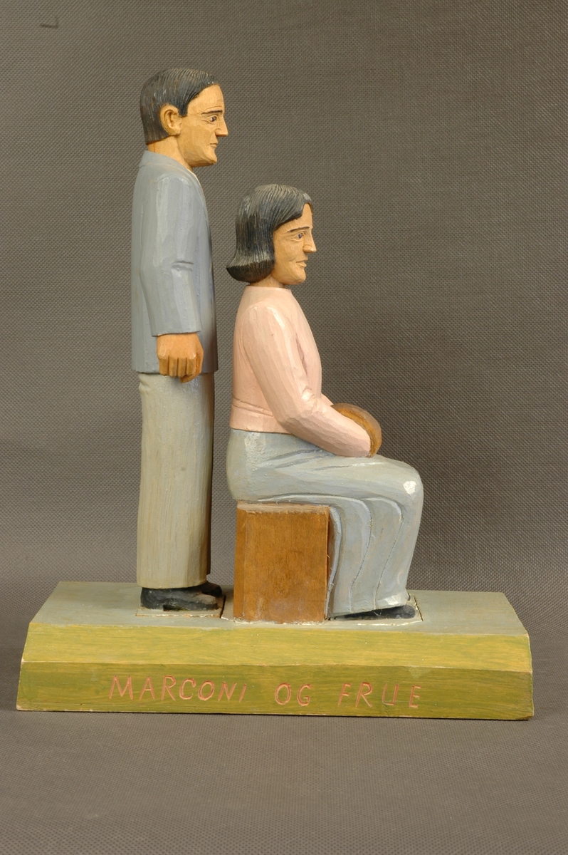 Skåret og delvis malt trefigur på sokkel. Figuren består av en mann og en kvinne. Begge personfigurene er løst nedfelt i sokkelen.