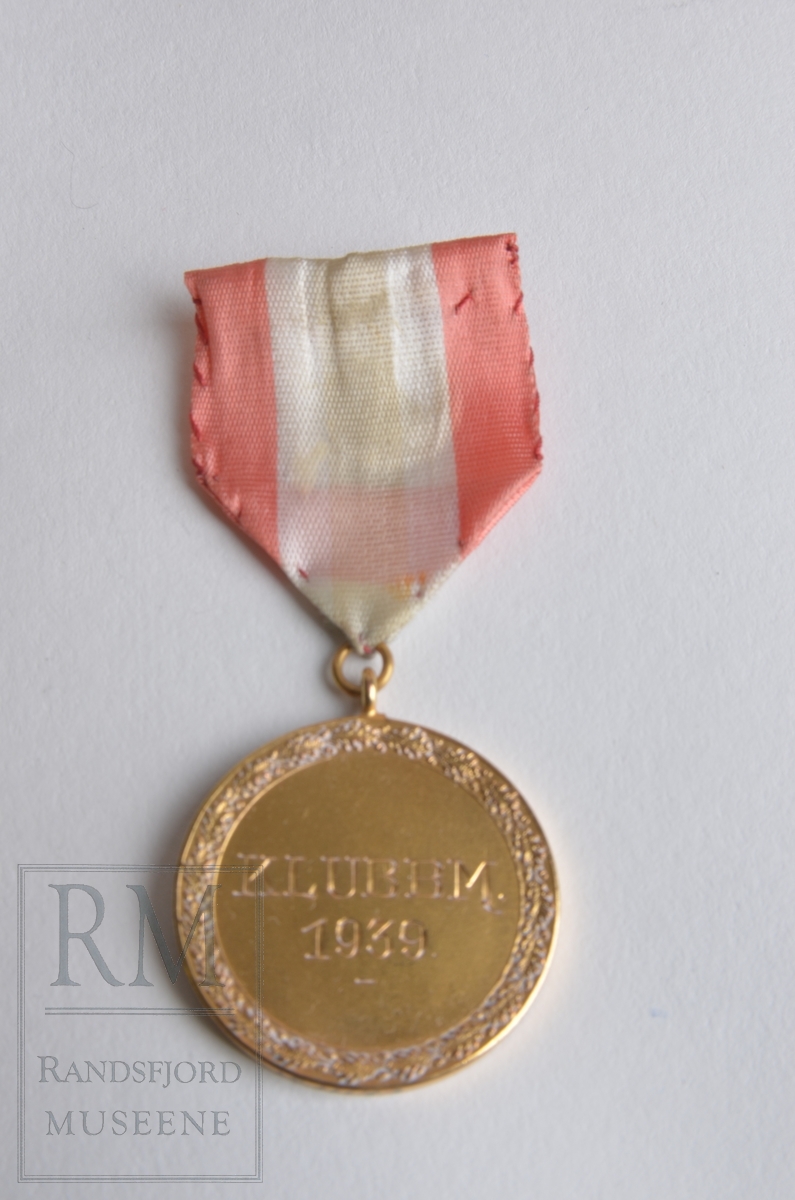 Gullmedalje. Gult metall. Ekebladkrans og inngravert: KLUBBM 1939. Baksiden slett. Medaljen henger i bånd i rødt, hvitt og blått (falmet) Påsydd nål på baksiden av båndet.