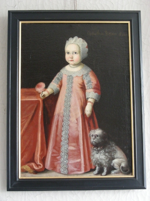 Barnporträtt, helfigur. Fotsid rosa klänning med spetsgarneringar. Mössa med spetsgarneringar.Bredvid sig  har flickan en liten sittande grå hund. På andra sidan skymtar ett bord med duk. Flickan håller i sin vänstra hand en rosa ros.