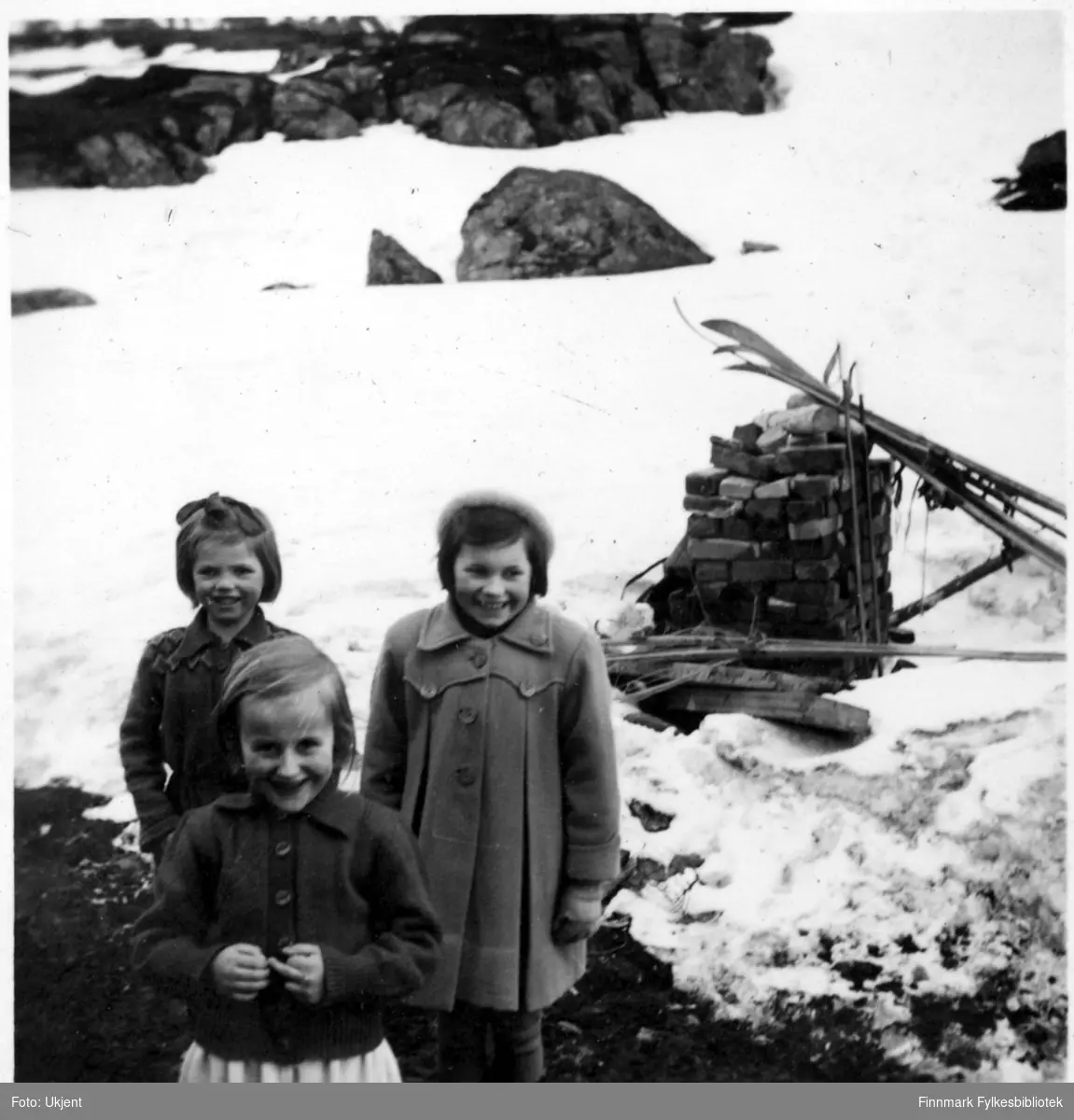 Jerlaug og Karin Andersen står bak Randi Martinsen (framst) og smiler til kameraet. Alle barna er fra Nuvsvåg. Jerlaug og Karin er kledd i jakker. Jerlaug har en sløyfe i håret, Karin en lue på hodet. Randi har på seg en genser. I bakgrunnen kan man se ski som er stablet opp, steiner og mye snø. Bildet er mulignes tatt i 1952.