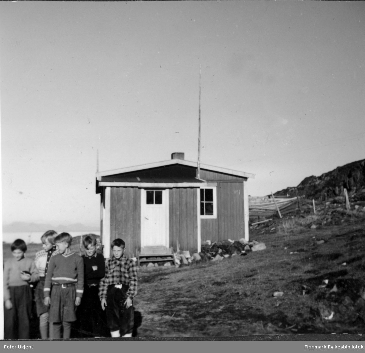 Elever ved skolen i Nuvsvåg 1952. Fra venstre: Per Nilsen, Henning Pettersen, Finn Rydheim, Evald Frisk og Adler Bårdsen. Guttene er kledd i knebukser, bukser, gensere og belter. Huset i bakgrunnen er en lærerbolig. Man kan også se gjerder og havet i bakgrunnen.