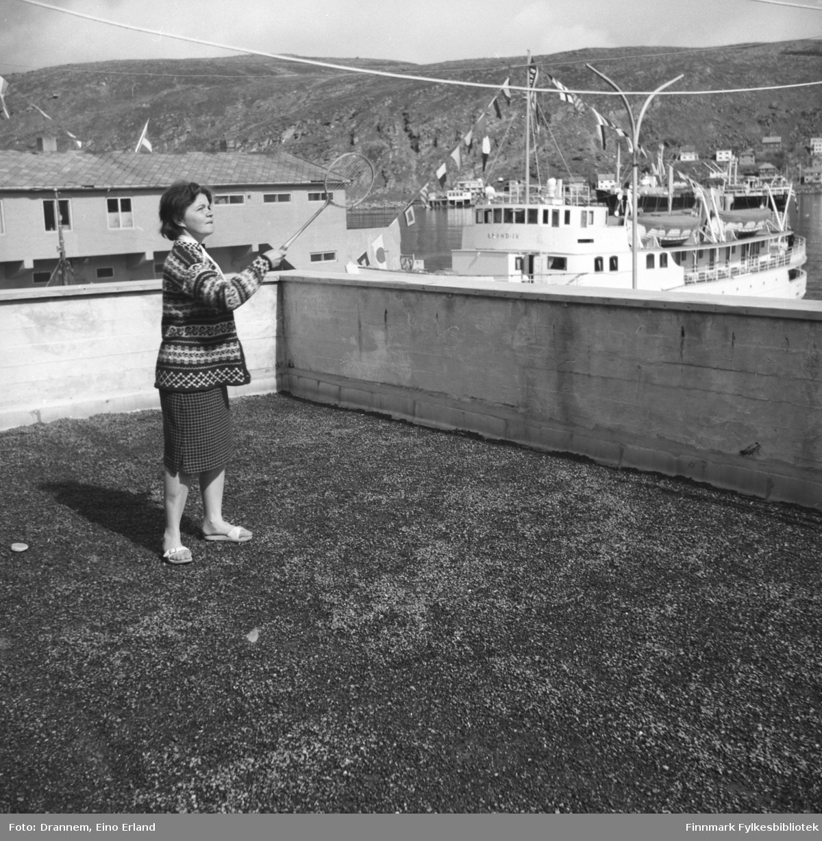 Maija Hoikka (født Gerasimoff) spiller badminton på verandataket til Tollboden. I bakgrunnen ved kai ligger båten Brand IV. Bak den kan man også se Vesterålskes godsbåt M/S Vardø på vei ut havna.
