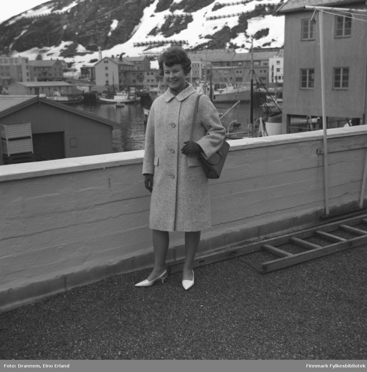 Turid Karikoski poserer for fotografen på verandataket til Tollboden i Hammerfest. I bakgrunnen ses indre havn med bebyggelsen langs Strandgata og fjellsiden til Salen.