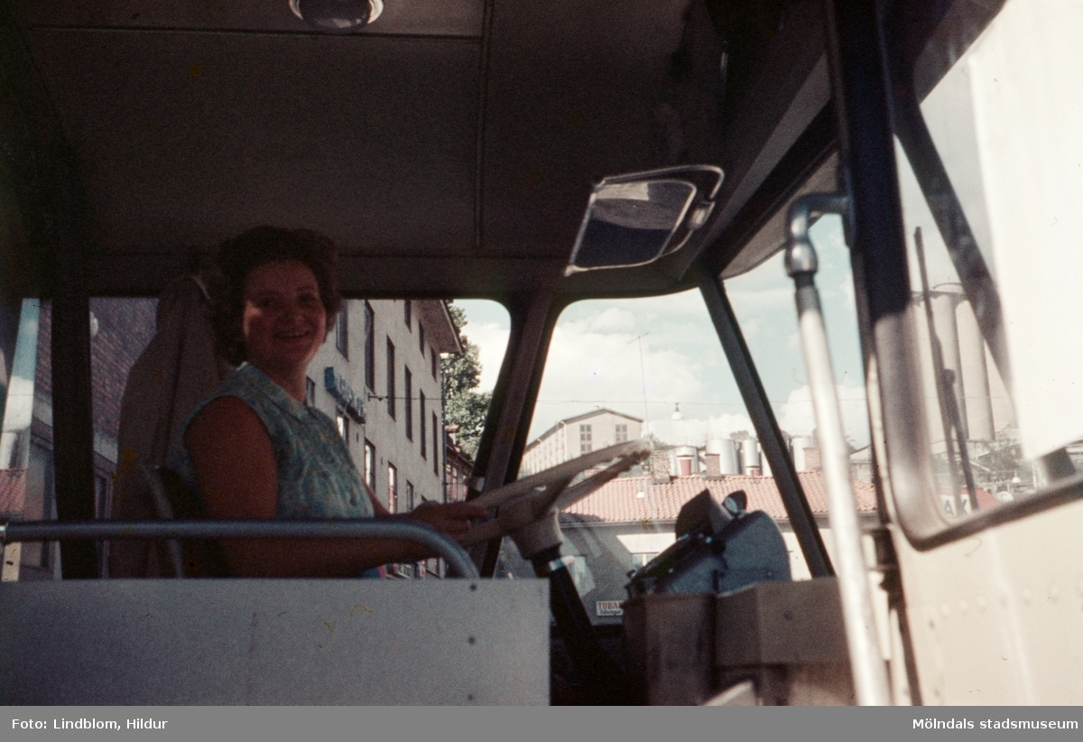 En kvinnlig busschaufför i sin buss vid Gamla Torget i Mölndal, 1970-tal. I bakgrunden, genom bussens rutor, skymtas husen från vänster Kvarnbygatan 41, 43 samt taket på nr 45.

För mer information om bilden se under tilläggsinformation.