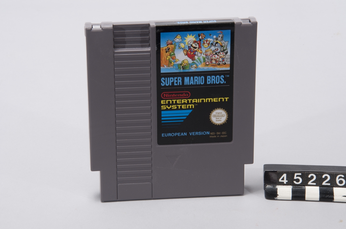 Cartridge med tv-spelet Super Mario Bro's inklusive kartong.  Super Mario Bros var ett sidscrollande plattformsspel, något som infördes i och med spelkonsolen NES vilket gjorde att spelvärlden kändes nästintill oändlig i jämförelse med de statiska spelplaner som tidigare spelkonsoler kunde hantera.  Spelet bestod av 8 världar med 4 banor i var värld.