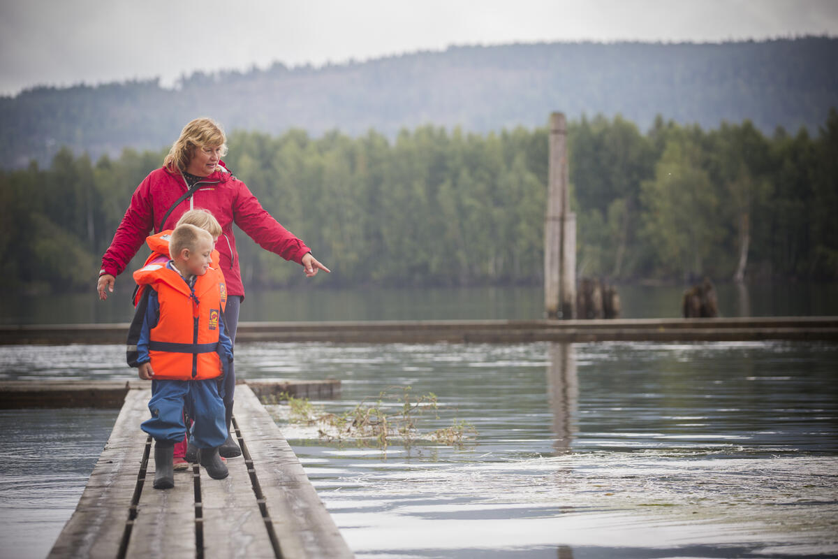 En voksen dame og to små barn går på flåteganer i elva. Begge barna har oransje redningsvester på seg. (Foto/Photo)