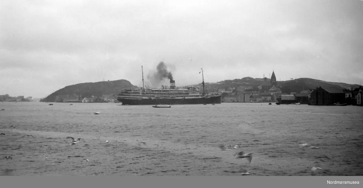 Turistbåten på bildet er franske M/S Foucauld på 11028 tonn, bygget for rederiet Compagnie des Chargeurs Reunis i 1923 som
kombinert laste- og passasjerskip D/S L'Hoedic. Skipet ble ombygget til dieseldrevet turistskip i 1929 og gitt navnet M/S Foucauld.
Skipet gikk tapt 15.juni 1940 i La Pallice etter et tysk flyangrep. Se også bildenr. KMb-1989-017.0037 (Info fra Espen Arnestad - 04.03.2017) -  Ute i havnebasenget i Kristiansund ser vi et passasjerskip, med Nordlandet og Nordlandet kirke i bakgrunnen.  Vi ser også litt av Innlandet stikkende ut helt til høyre i bildet. Fra Nordmøre Museum sin fotosamling. Serie