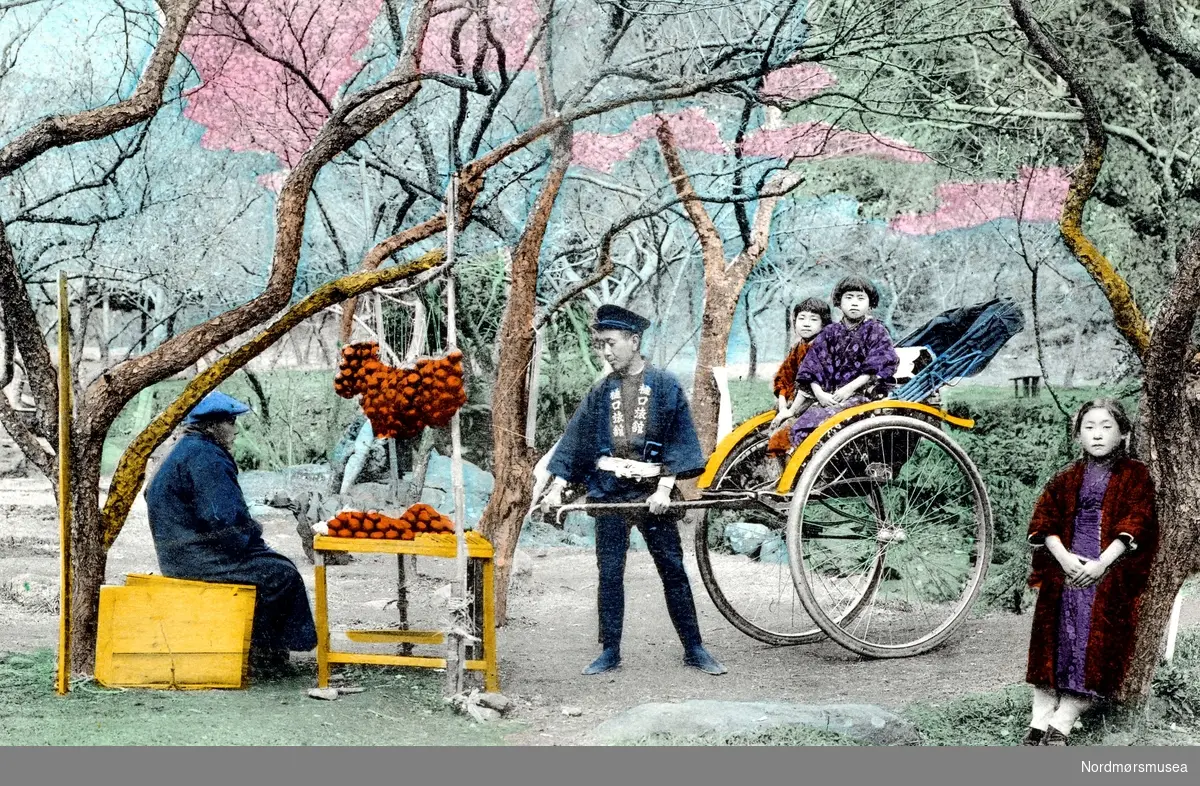 Kolorert postkort, trolig fra Japan, hvor vi ser en rickshaw med to gutter ombord, en kvinne like ved og en mann som trekker vognen. Like ved ser vi også et mann sittende ved sitt torgutsalg. Datering og fotograf er ukjent. Fra Nordmøre Museums fotosamlinger. /Reg:EFR2013/
