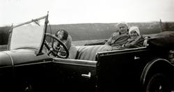 Foto av en mann og to kvinner, sittende i en bil som står pa