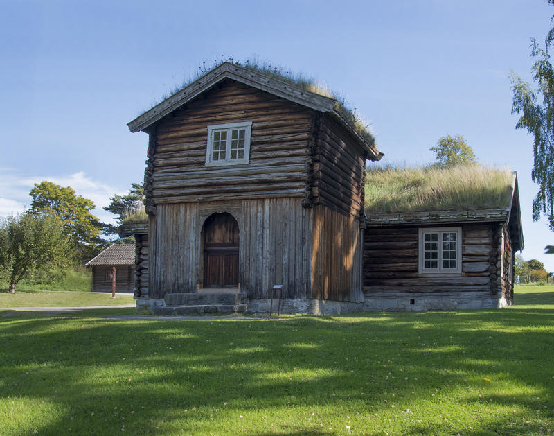 Lav, grå tømmerbygning med gress på taket og toetasjers tilbygg, et såkalt "barfrø" midt på langveggen.