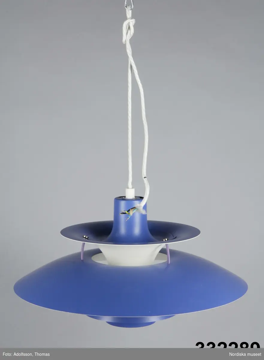 Taklampa, elektrisk, s.k. PH-lampa, uppbyggd av ett system av plåtskärmar, fyra yttre och två mindre skärmar inuti lampan. 
Högst upp behållare för glödlampans sockel monterad i en trumpetformad övre skärm med öppningen uppåt. Under och omkring denna finns lampans vidaste skärm, en tallrik med 50 cm i diameter. Lampan avslutas nedåt av tre mindre skärmar, underskärmen nästan helt cylindrisk. Skärmarna hålls ihop av tre vertikala metallstag.

Tampan blålackerad, synliga inre skärmar vitmålade, skärmar dolda för ögat rödmålade. 

Textilklädd sladd utan stickpropp, lampan har varit fast monterad.
/Maria Maxén 2014-01-27