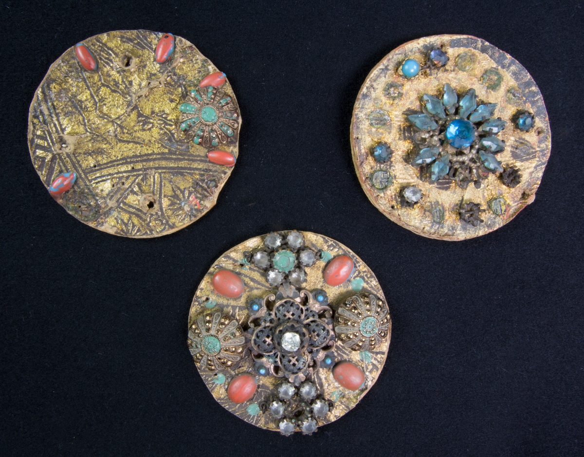 Smycke, 3 st, runda av guldmålad papp med glasbitar, knappar med mera fästade med metalltenar samt klistrade på pappskivorna.