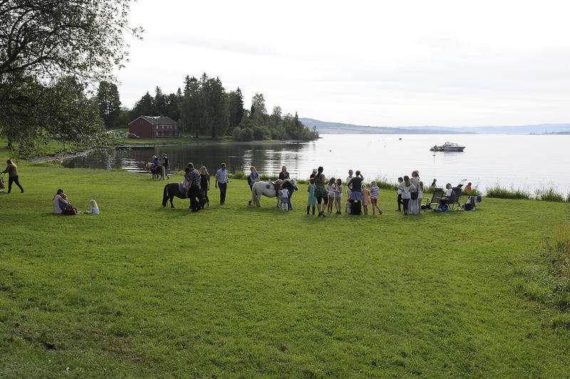 Barn og ungdom som driver med aktivitet på grønn gresslette ved vannet, museumshus i bakgrunnen.