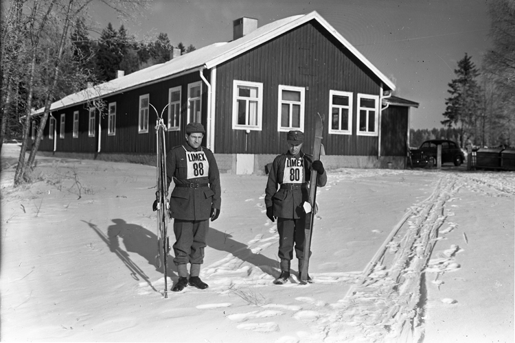 Två skidlöpare står framför en barack en vinterdag.
I bakgrunden syns en bil.
"Limex - skidfabriken Limex i Tobo bruk, Tegelsmora socken,
Uppland.