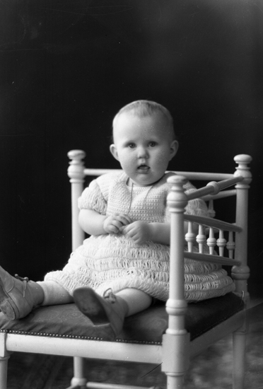 En liten flicka i vit klänning sitter på en stol.
Trol. dottern till familjen Petersson på MINI00007. 
Ateljéfoto.
Rut Signe Marianne Petersson (1934-   ), Hjärtenholm.
Källa: Församlingsbok, Lekaryd 1924-1941.