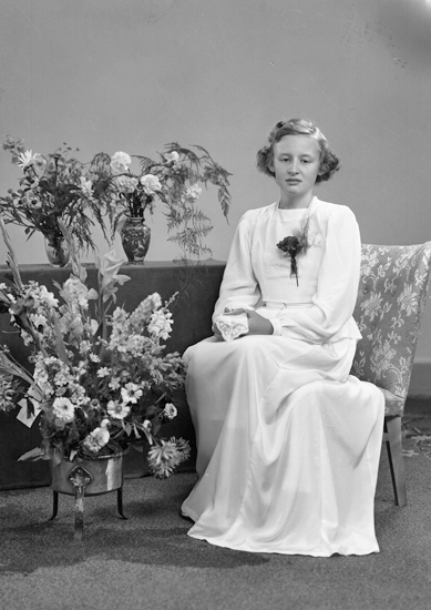 Foto av en kvinnlig konfirmand i vit konfirmationsklänning. I händerna håller hon en spetsnäsduk. Hon sitter på en stoppad stol, omgiven av blommor.
Helfigur. Ateljéfoto.