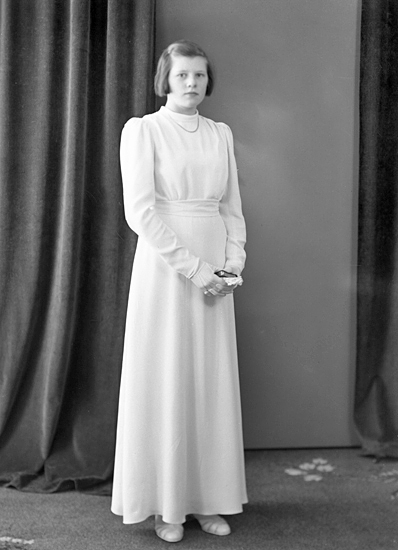Foto av en kvinnlig konfirmand i lång, vit konfirmationsklänning. I händerna har hon en liten psalmbok.
Helfigur. Ateljéfoto.