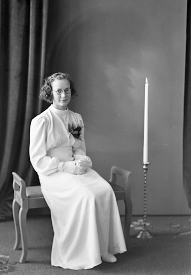 Foto av en kvinnlig konfirmand i lång, vit konfirmationsklänning. Hon sitter på en stoppad bänk, med en stor golvljusstake bredvid.
Helfigur. Ateljéfoto.