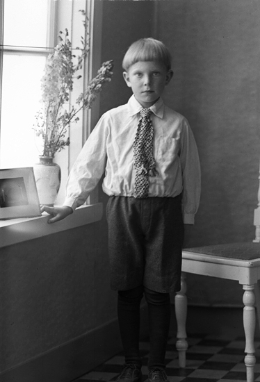 Foto av en pojke i kortbyxor, skjorta och slips som står vid ett fönster. Bredvid honom syns en vitmålad stol.
Helfigur. Ateljéfoto.