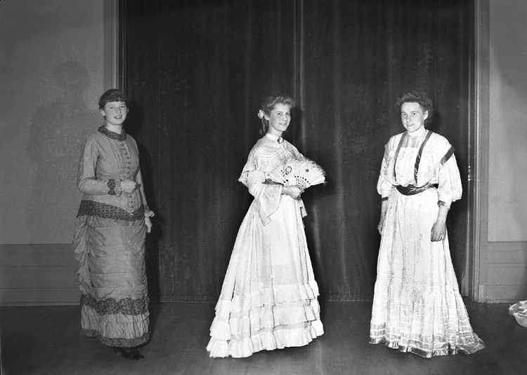 Tre kvinnor.
Till vänster syns en kvinna i klänning från 1870-talet , i mitten från 1850-talet  och till höger en kvinna i klänning från ca 1900.