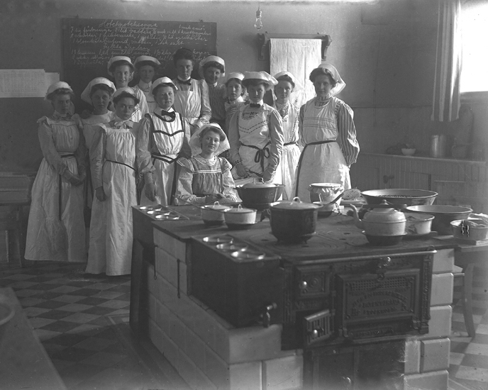 Foto av en grupp unga kvinnor iförda stora förkläden och hättor,  i ett skolkök.
