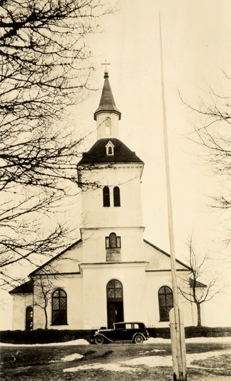 Foto på en kyrka framifrån med en bil och en flaggstång utanför.