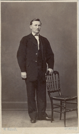 Porträtt (helfigur, en face/halvprofil) av en stående man i mörk kostym med vit skjorta m.m. Han håller ena handen på ryggstödet till en svarvad stol. 

Otto Hellberg - (1845-1904), sjökapten, Uddevalla, f. i Grinneröd.
Källa: Bl a Folkräkning 1880, Uddevalla stad.
