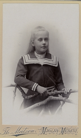 Porträtt (knäbild), ateljé, av en ung flicka i sjömansklänning, stående vid en konstgjord gärdesgård.