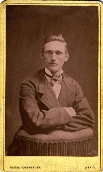 Porträtt (bröstbild, en face) av en okänd sittande man, klädd i stärkkrage, fluga och mörk kavaj. 
Han sitter bakvänt på en stoppad stol och vilar armbågarna mot ryggstödet.
