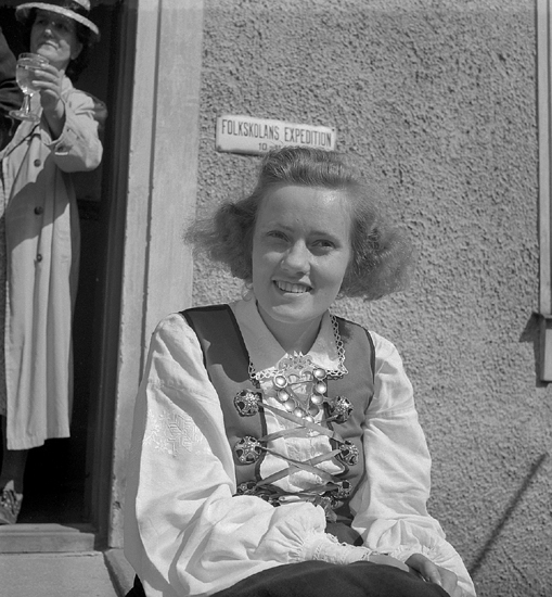 Barnens Dag, 31/5 1946.
En ung kvinna klädd i Värendsdräkt, sitter i solen på trappan till Folkskolans expedition. Bakom henne skymtar en äldre dam i kappa och hatt, som har ett glas i handen.
