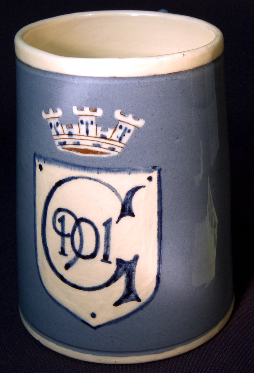 Ölsejdel av keramik. Utsidan glaserad och försedd med vapensköld i vitt med mörkt blå text: G 1901. Insidan vitglaserad. Över skölden en krona. Tillverkad som souvernir för Gävleutställningen 1901.