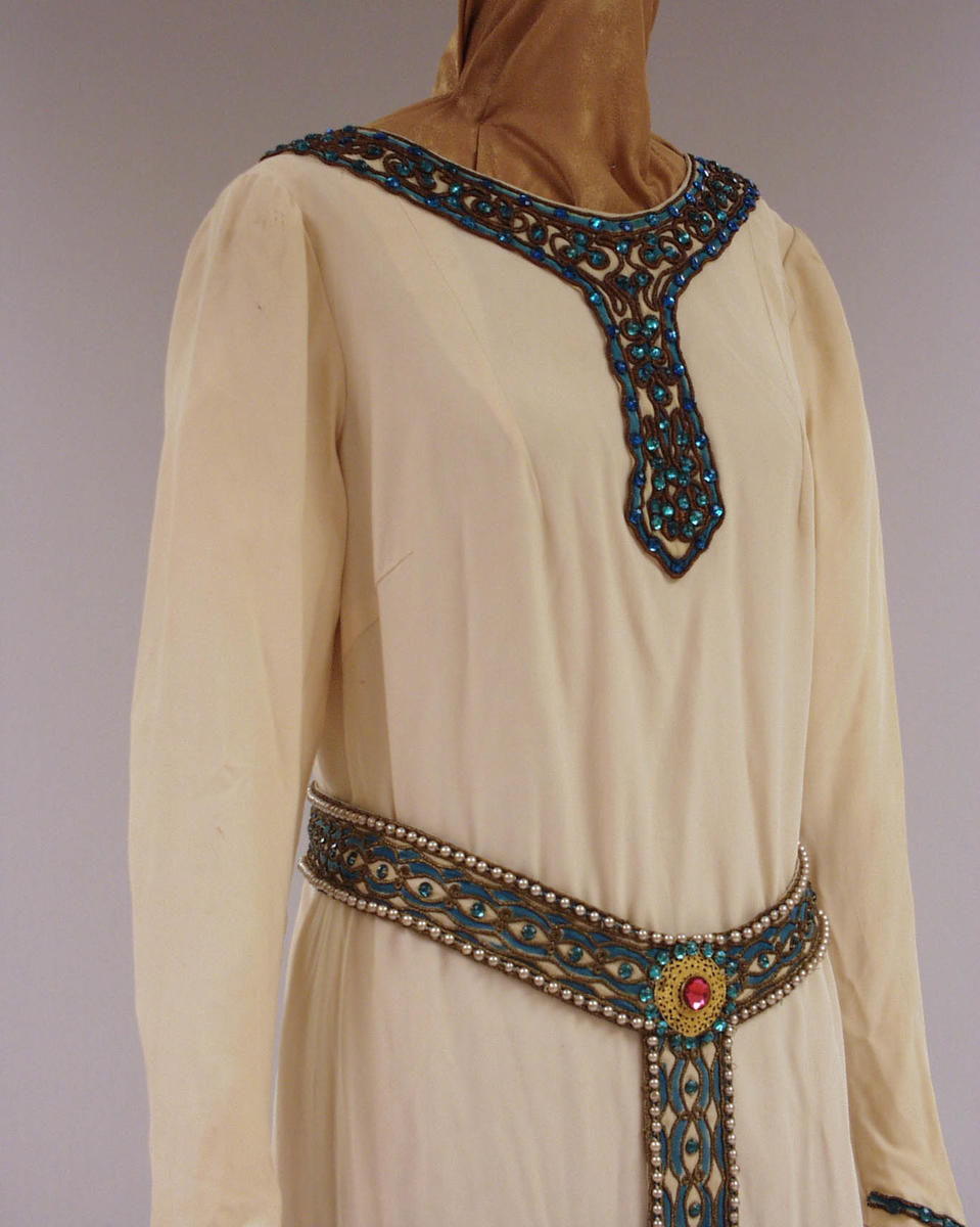 Kostyme Kirsten Flagstad i rollen som Elisabeth i Tannhauser. Hvit kjole med detaljer av perler i halsen og nedover brystet. Matchende belte rundt livet.