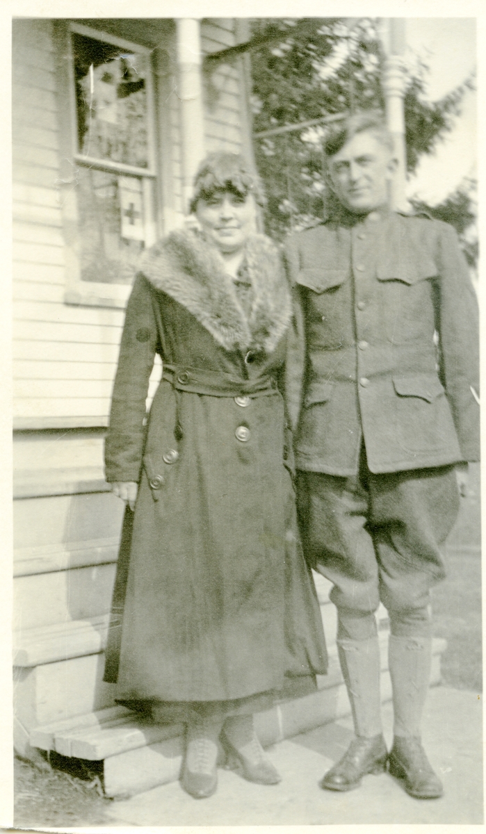 Mann og kvinne avbildet utenfor et hus. Han er kleded i en uniform og hun har en lang mørk kåpe med skinnbestetning.