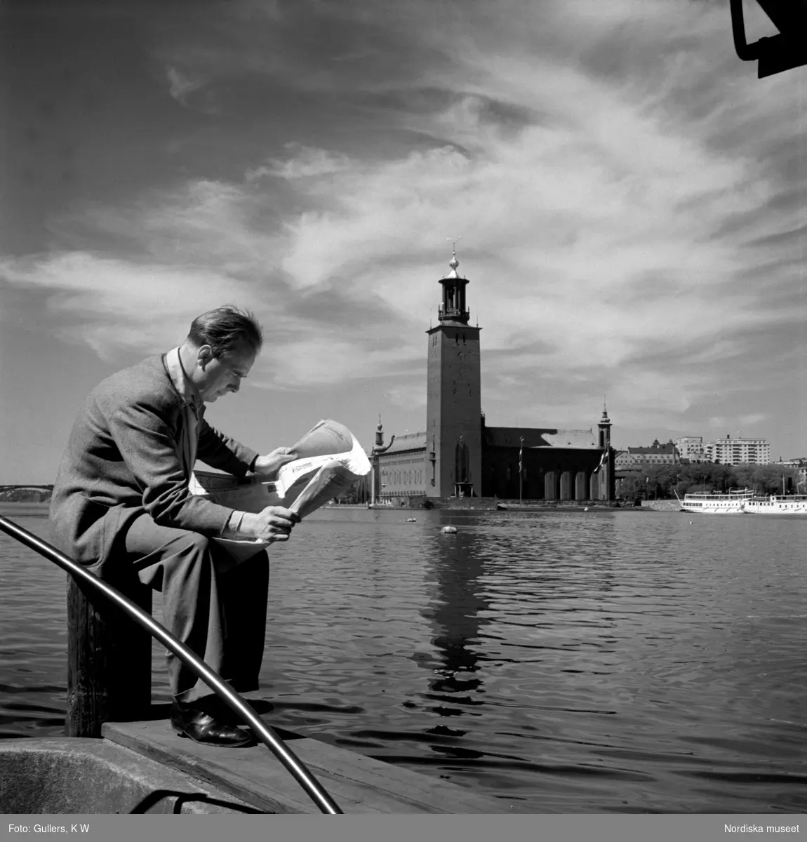 En man sitter på en pollare och läser en tidning. Stockholms stadshus och Riddarfjärden i bakgrunden.