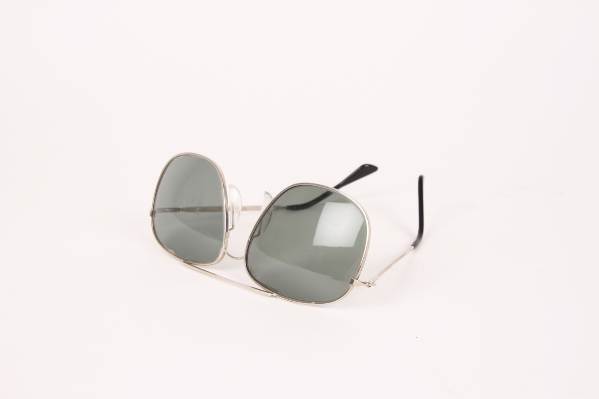Et par solbriller med metallinnfatning. Brillens modell er pilotbrille, med sølvfarget innfatning og svarte glass. Svart plast på enden av hver brillestang.
