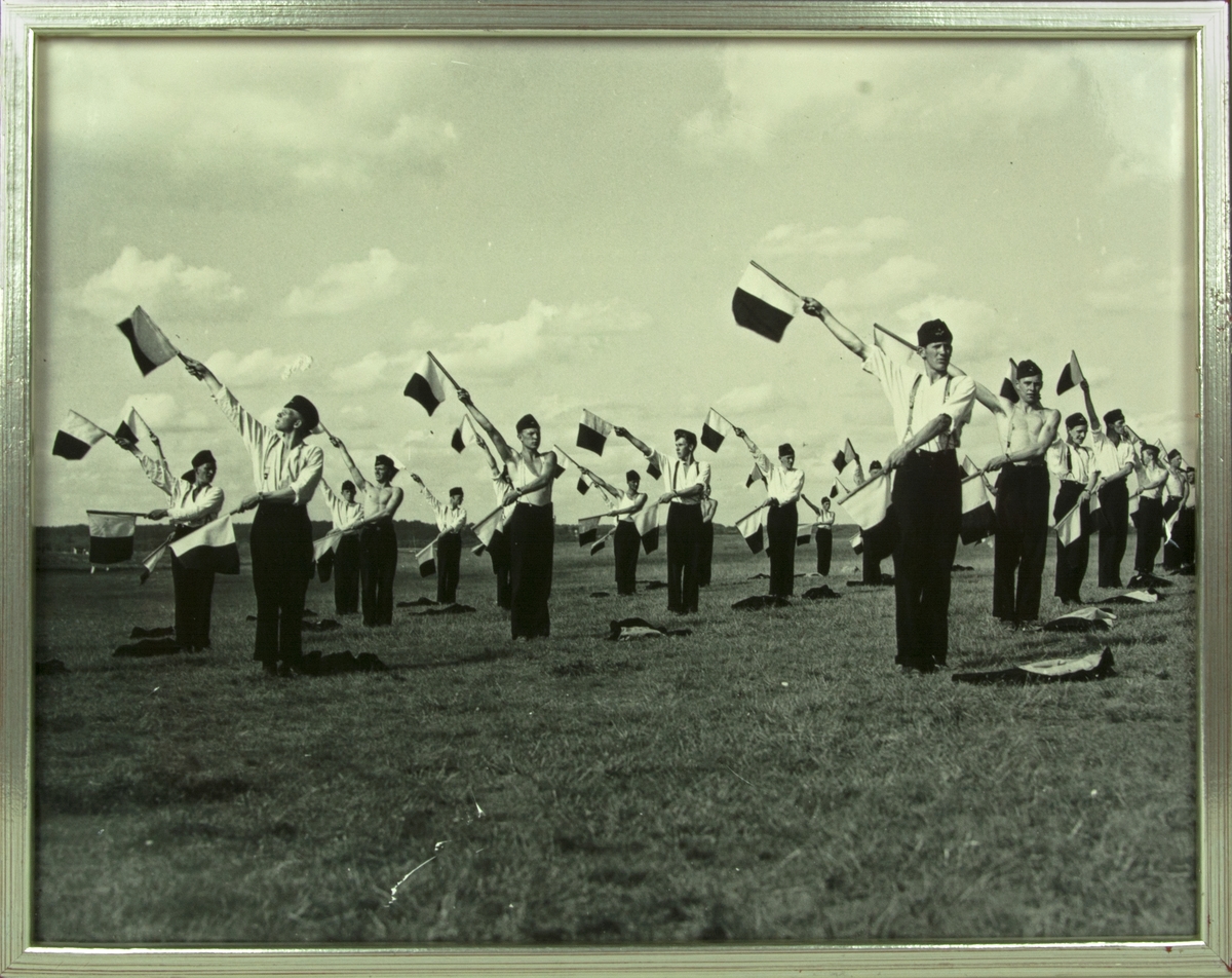 Signalering ASP-AVD på F 5 i Ljungbyhed, 1937. Värnpliktiga män övar signalering med flaggor mot flygplan. Inramat foto.