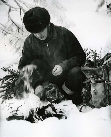 En man sitter på en hög med granris i snön. Han har gjort upp en liten eld. Bredvid honom i snön står hans öppna ryggsäck.