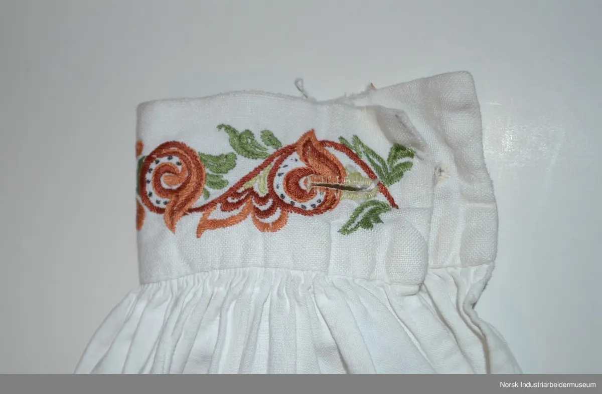 Plattsøm på kraven, mansjettene og bringa. Skjorten er åpen foran og helt ned.