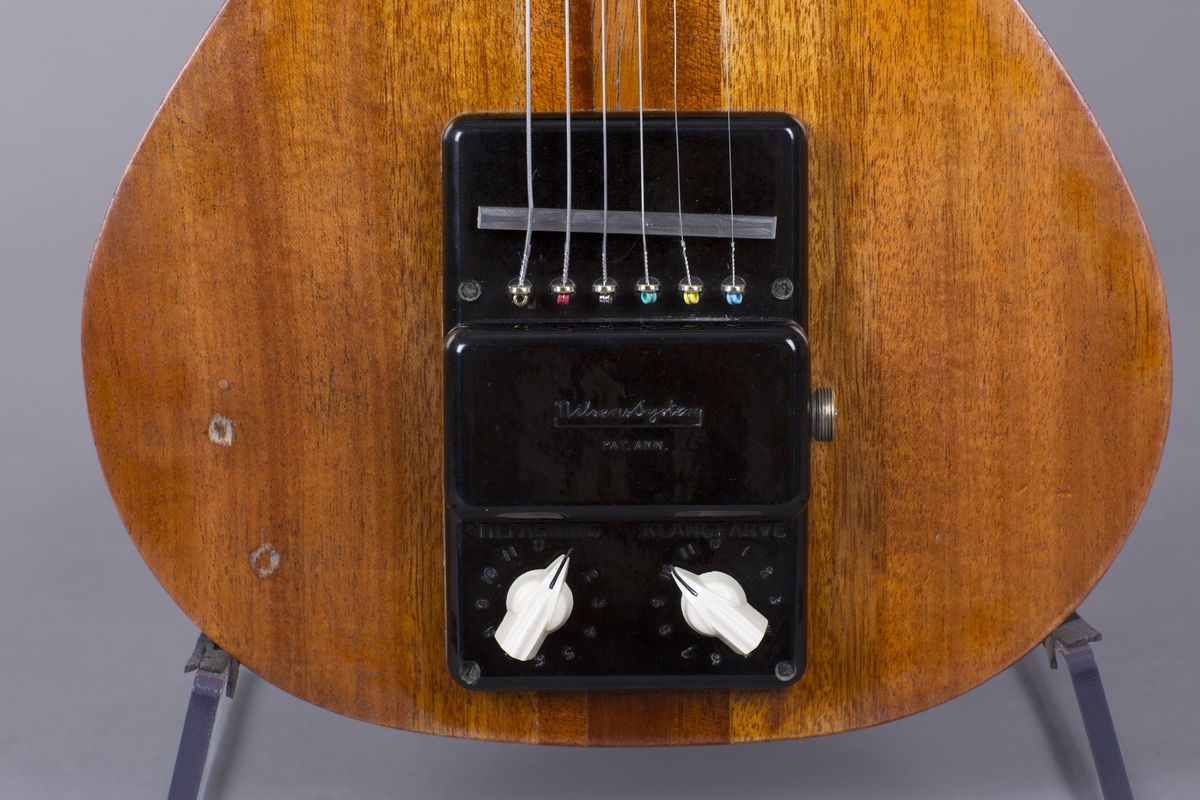 Elektrisk gitar med kropp og hals av mahogny. Magnet ved halsfeste og pickup ved strengefeste (NilsensSystem). Kontrollere for tilpasning og klangfarge. Tilpasning tilsvarer volum og gir størst volum på nivå 1. Enkelte av stemmeskruene er ikke originale.