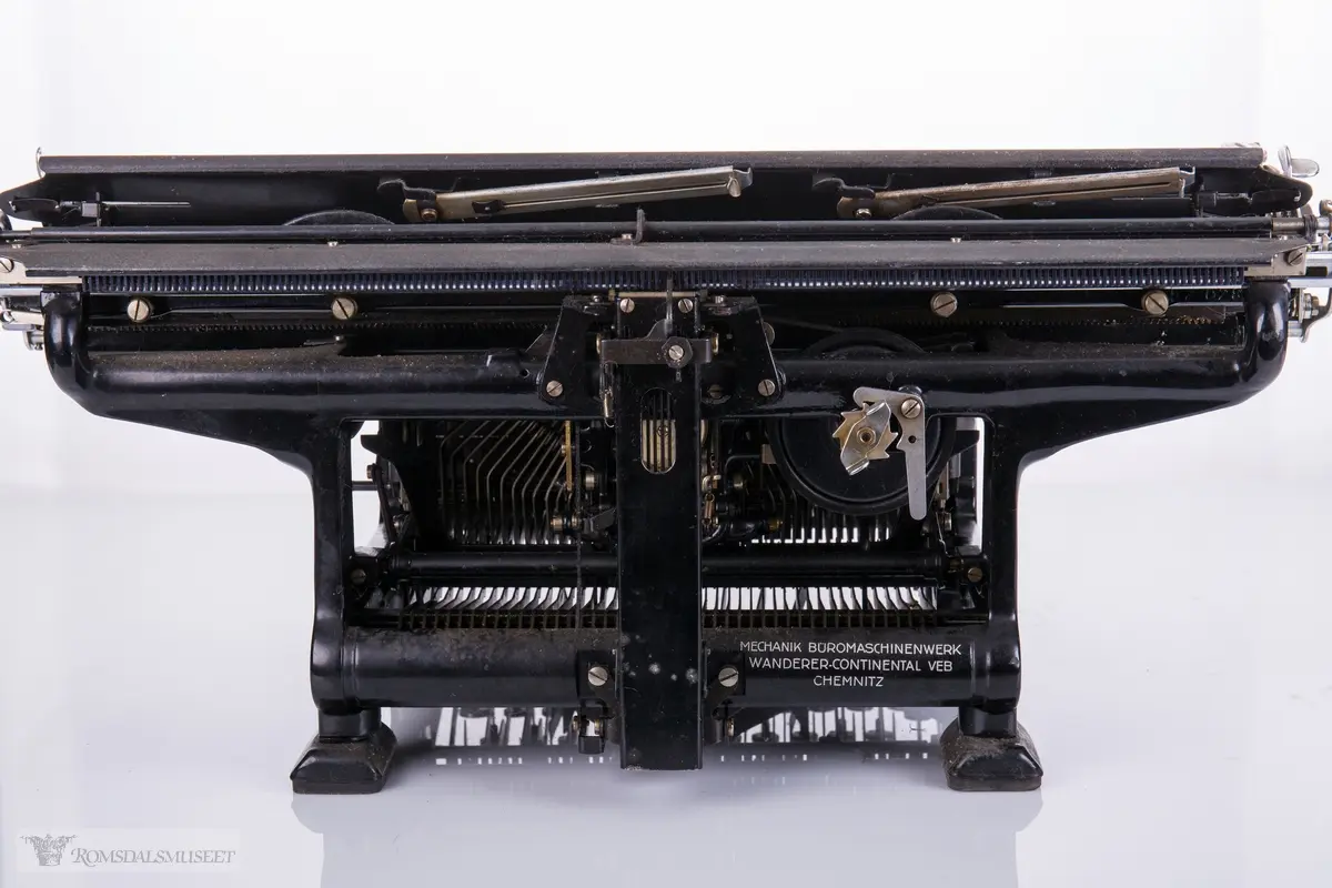 Rektangulær, mekanisk skrivemaskin på fire bein med en bred overdel som går langt utover maskinens grunnform. 
56 tangenter.