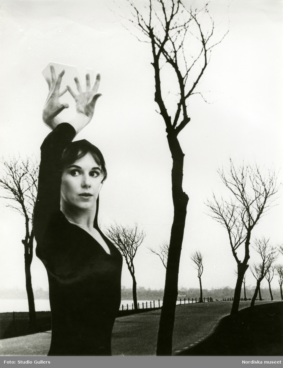 Kvinna poserar med uppsträckta armar framför en landskapsbild med väg och kala träd, troligen montage