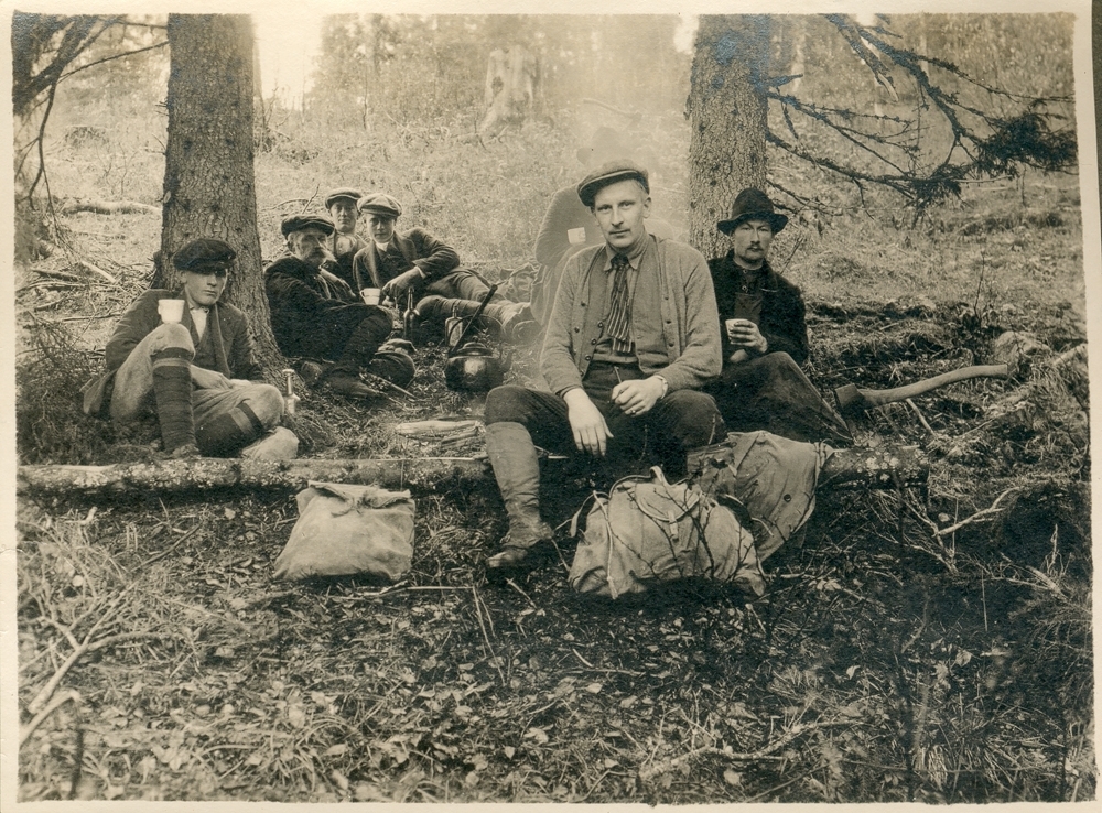 Utendørs. Gruppe. En gruppe menn sitter ute i skogen og drikker kaffe, slapper av eller spiser. Matpause. De har sekker foran seg. De ryker fra et bål med en kaffekjele over. Arbeidere som jobber med stikning av kraftlinjen nordover til Storforshei og sydover til Gullsmedvik (DIOC).
