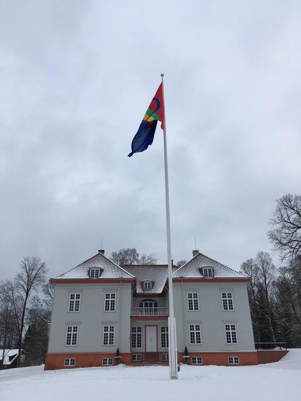 Vi ser vest-fasaden (hoved fasaden) på Eidsvollsbygningen og det samiske flagget vaier i vinden i hovedflaggstanga foran bygningen. På bakken og taket er det snø.